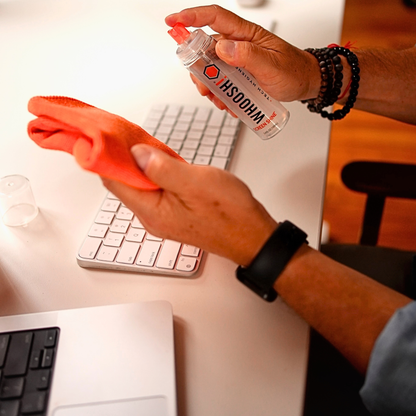 Wiping a Mac keyboard with the orange Whoosh microfiber cloth while holding the Go XL bottle