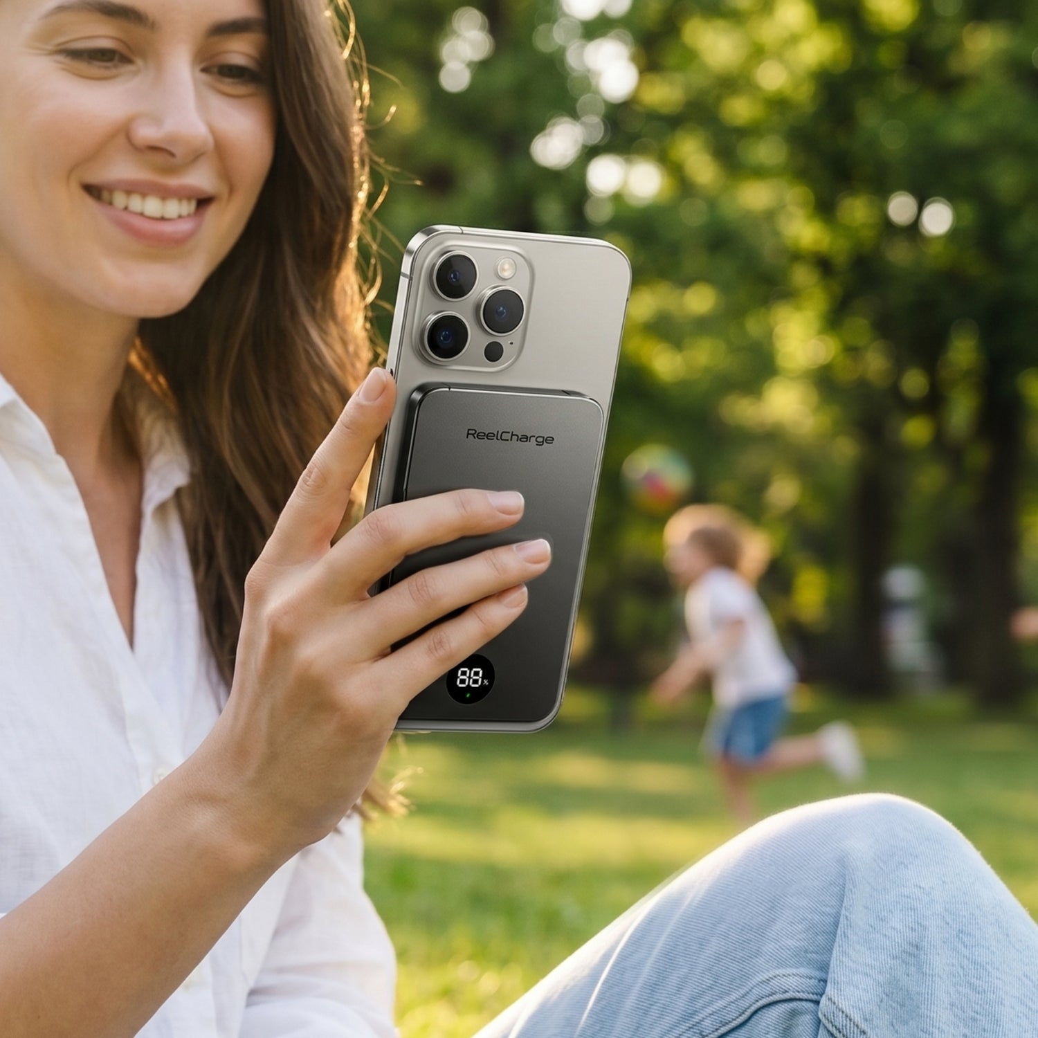 Woman in a park holding an iPhone with the gray ReelSafe 5,000 mAh power bank attached to the back